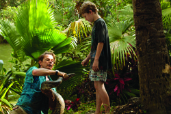 A man in a teal shirt holds a large snake while a young boy stands nearby in a lush, tropical garden.