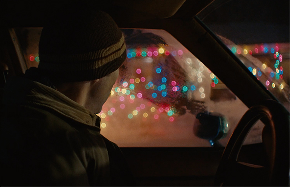 View from inside a car of a person in a beanie looking out at blurry red, blue, and green lights.