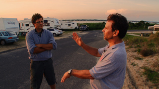 Two men stand on a paved road near parked RVs and cars during sunset.