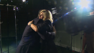 A woman in a dark outfit embraces a man in a dark suit under bright studio lights.