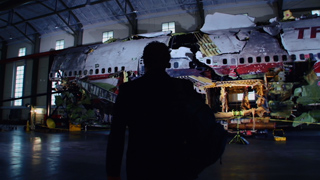 Silhouette of a person facing a large, partially dismantled airplane fuselage inside a dimly lit hangar.