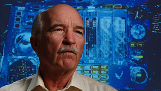 A bald man with a mustache looking thoughtfully to the side against a blue background of technical diagrams.