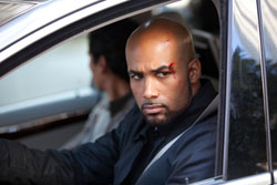 A bald man with a small cut on his cheek looks intensely toward the camera while sitting in a vehicle.
