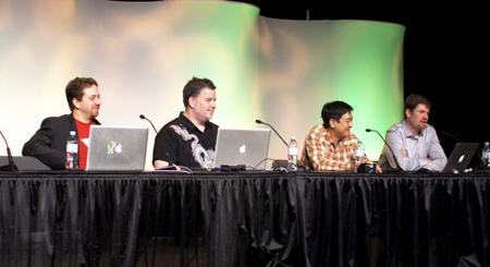 Four men sit behind a long table with laptops and microphones against a colorful backdrop.