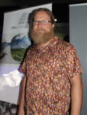 Man with glasses and a long beard wearing a patterned shirt stands in front of a banner.